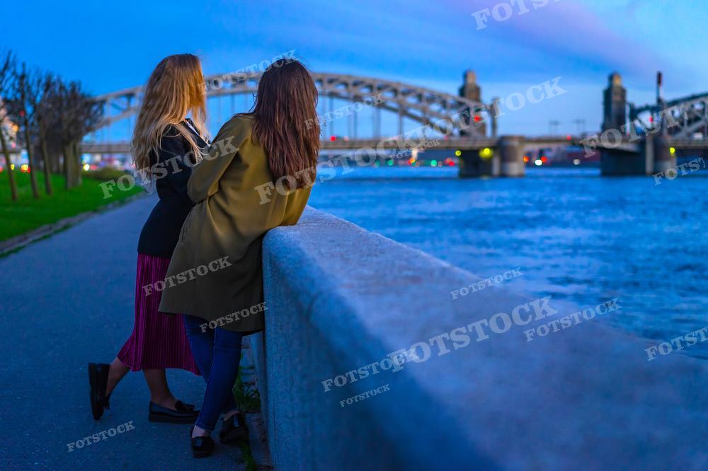 На фотографии изображены две женщины, стоящие на набережной и смотрящие на реку в вечернее время. Они облокотились о перила, погруженные в разговор или наслаждающиеся видом. На заднем плане виден один из знаменитых мостов Санкт-Петербурга, мягко освещенный и отражающийся в водах реки. Окружающее освещение и голубоватый оттенок неба указывают на то, что время суток близко к сумеркам. Это изображение может быть использовано в тематических статьях о городской жизни, путеводителях по Санкт-Петербургу или в качестве визуального материала для социальных сетей и блогов, подчеркивающего атмосферу города.